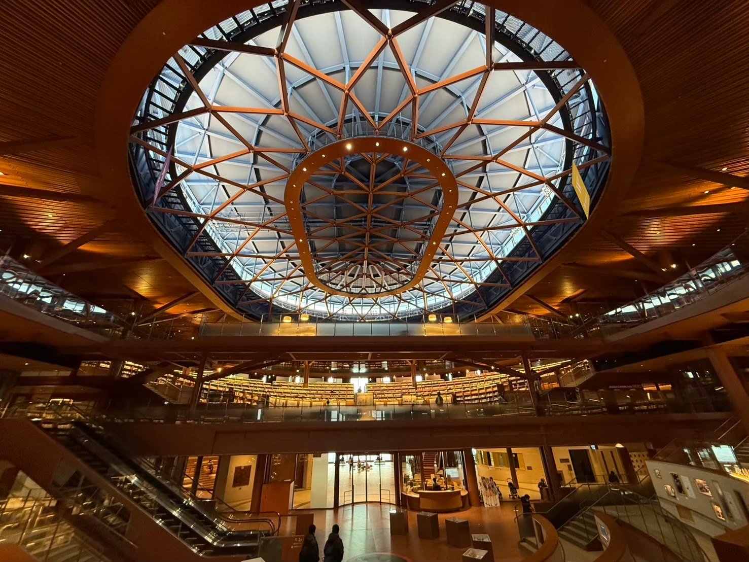 The domed atrium of Ishikawa Prefectural Library, with tiered bookshelves ringing the central hall under a wooden lattice skylight — 石川県立図書館の吹き抜け。木格子のドーム天井の下に、円形に配置された書架が段状に並ぶ。